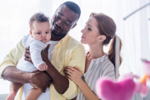 A joyful family of three, with a father holding a baby and a mother beside them, all smiling in a bright, warm setting.