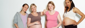 Four pregnant women, smiling and standing together, each showcasing their baby bumps, in casual attire against a light background.