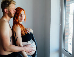 A couple embraces by a window, with the woman showing her baby bump, highlighting a moment of intimacy and anticipation.