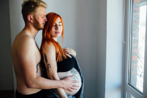 A couple stands together by a window, with the woman cradling her pregnant belly while the man embraces her from behind.