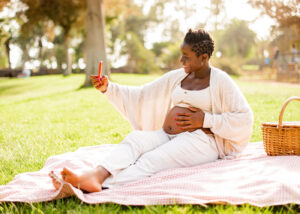 A pregnant woman sits on a blanket in the park, taking a selfie while gently touching her belly, surrounded by greenery.