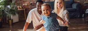A joyful toddler in casual attire stands with smiling parents in a cozy, well-lit room featuring brick walls and plants.
