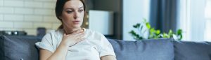 A woman appears anxious while sitting on a couch, holding her throat, with a pensive expression in a cozy living room.