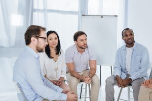 A group of five people in a seated discussion, with light-colored clothing and a blank whiteboard in the background.