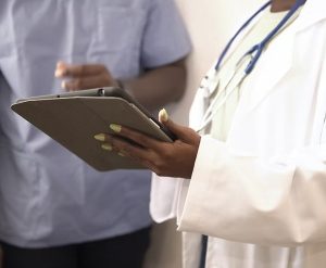 A person in a white coat holds a tablet, while another individual in scrubs stands nearby, suggesting a medical setting.