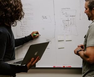 Two individuals discuss ideas at a whiteboard, one holding a laptop and the other writing notes with a green marker.