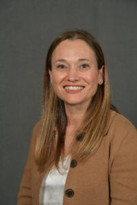 A woman with long hair, wearing a tan jacket, smiles warmly against a gray background.
