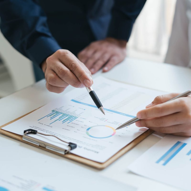 Two people discussing data on a clipboard with charts and graphs, using pens to point at specific details.