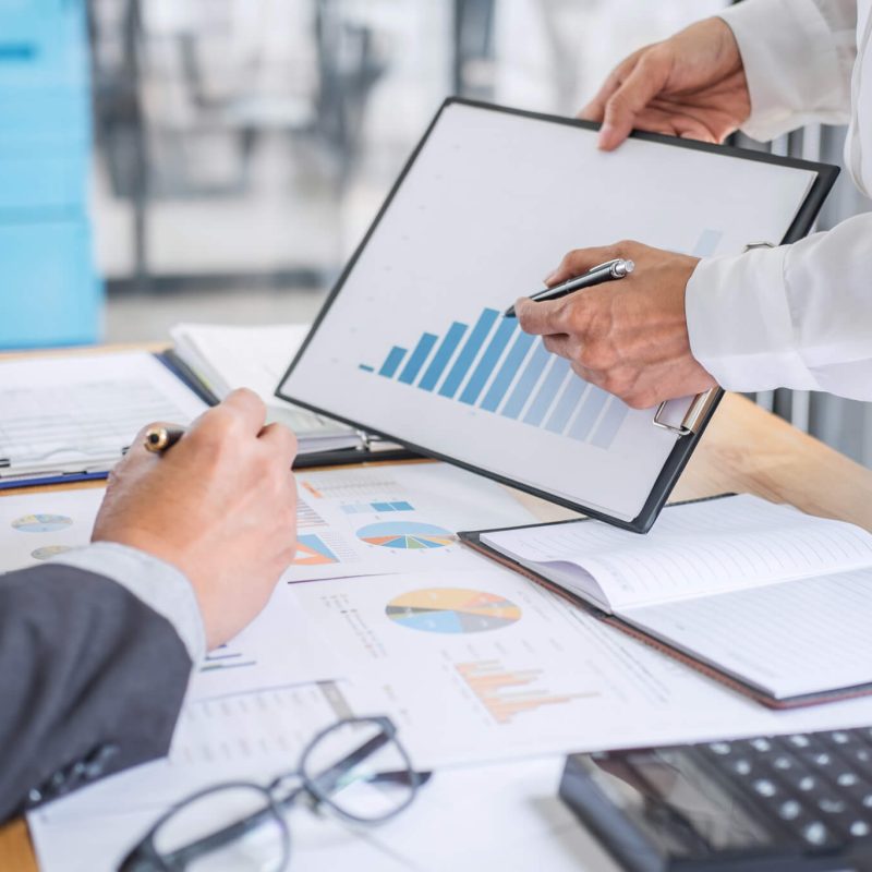 Business professionals reviewing charts and graphs on a tablet during a meeting, surrounded by documents and a calculator on a wooden table.