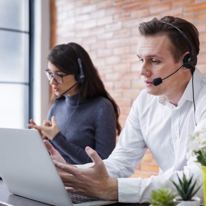 Two people in a modern office setting, engaged in a call, using laptops and headsets, with plants and a brick wall in the background.