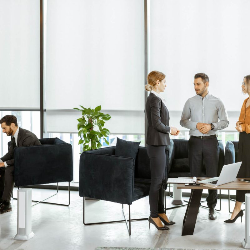A modern office environment with four professionals discussing, while one works on a laptop. Bright sunlight filters through large windows.