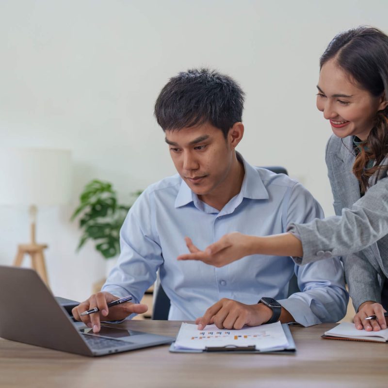 A focused young man works on a laptop while a smiling woman gestures, possibly offering guidance, in a modern office setting.