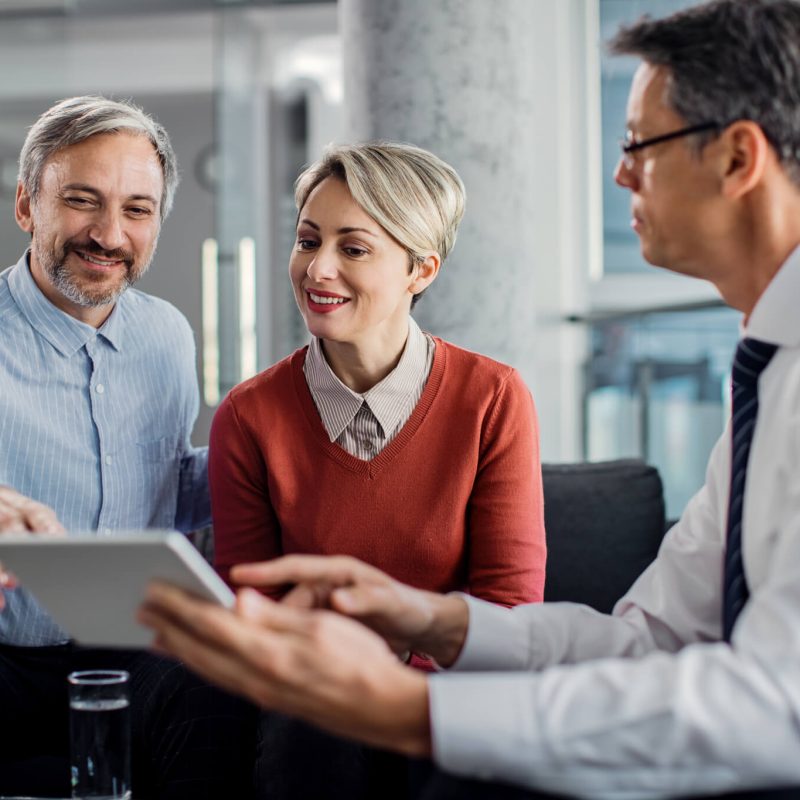 Three professionals engage in a discussion while reviewing information on a tablet in a modern office setting.