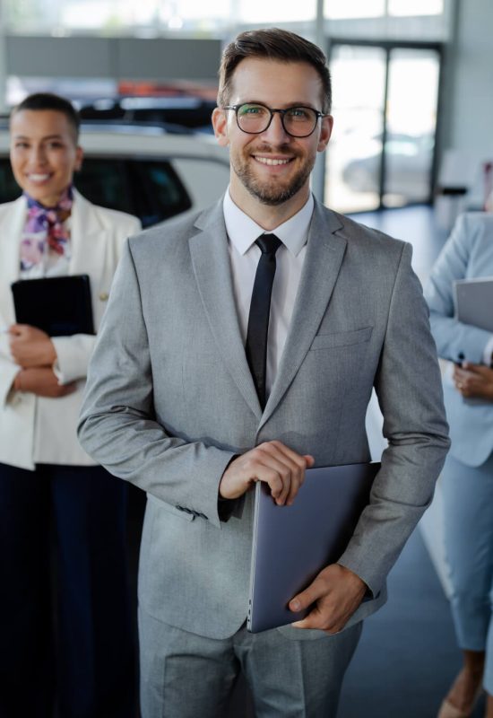 Three professionals in formal attire stand in a car dealership, smiling confidently while holding digital devices, surrounded by vehicles.